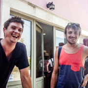 Deux amis souriants posant devant une boutique aux façades colorées en Crète, capturant un moment convivial et détendu.