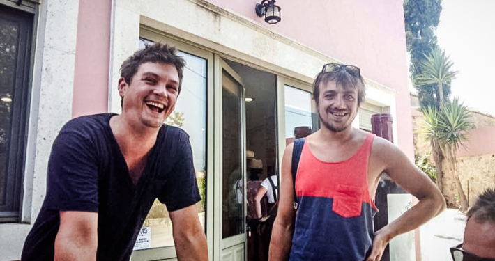 Deux amis souriants posant devant une boutique aux façades colorées en Crète, capturant un moment convivial et détendu.