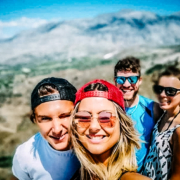 Groupe de quatre amis souriants prenant un selfie avec un paysage vallonné de Crète en arrière-plan, sous un ciel bleu éclatant.