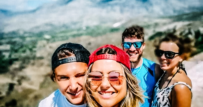 Groupe de quatre amis souriants prenant un selfie avec un paysage vallonné de Crète en arrière-plan, sous un ciel bleu éclatant.