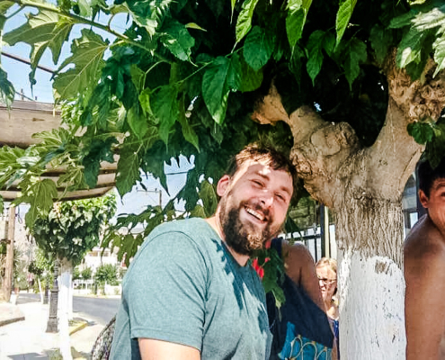 Groupe de personnes souriantes se tenant sous l’ombre d’un arbre en Crète, capturant un moment convivial et détendu dans un cadre ensoleillé.