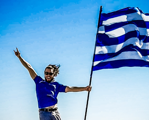 Homme joyeux tenant le drapeau grec flottant au vent sous un ciel bleu éclatant, symbolisant la fierté et la liberté en Crète.