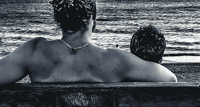 Vue arrière d’une femme et d’un enfant assis sur un banc face à la mer, contemplant l’horizon et une petite île au loin, capturant un moment paisible au bord de l’eau en Crète.