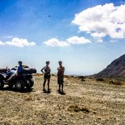 Trois personnes et un quad lors d’une pause dans les montagnes de Crète, sous un ciel bleu parsemé de nuages, avec une vue panoramique sur les paysages arides et les sommets environnants.