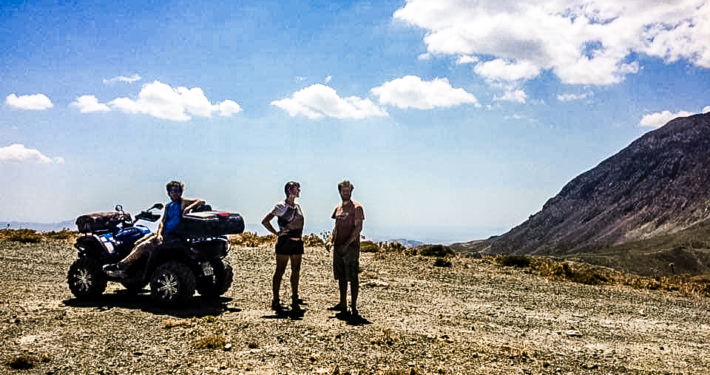 Trois personnes et un quad lors d’une pause dans les montagnes de Crète, sous un ciel bleu parsemé de nuages, avec une vue panoramique sur les paysages arides et les sommets environnants.