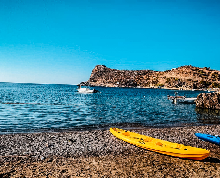 Plage ensoleillée en Crète avec des kayaks colorés sur le sable, des bateaux flottant sur une eau calme et des collines rocheuses en arrière-plan.