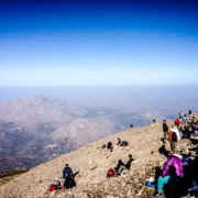 Groupe de randonneurs se reposant au sommet d’une montagne en Crète, avec une vue panoramique sur les chaînes montagneuses et un ciel bleu dégagé en arrière-plan.