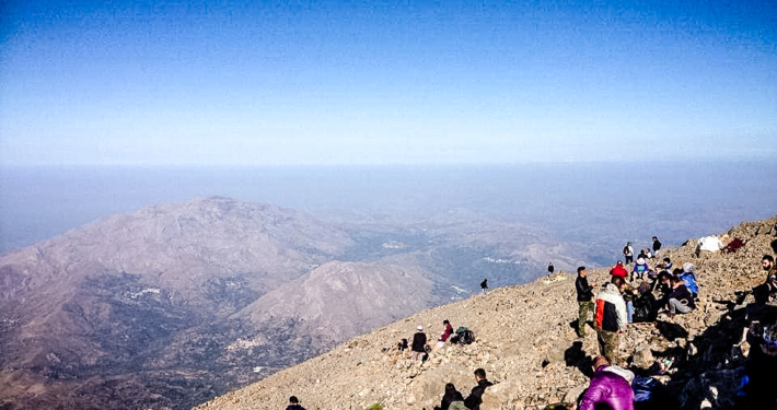 Groupe de randonneurs se reposant au sommet d’une montagne en Crète, avec une vue panoramique sur les chaînes montagneuses et un ciel bleu dégagé en arrière-plan.