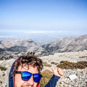 Manolis souriant prenant un selfie au sommet des montagnes en Crète, avec une vue panoramique impressionnante sur les vallées et les sommets environnants sous un ciel dégagé.