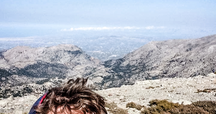 Manolis souriant prenant un selfie au sommet des montagnes en Crète, avec une vue panoramique impressionnante sur les vallées et les sommets environnants sous un ciel dégagé.