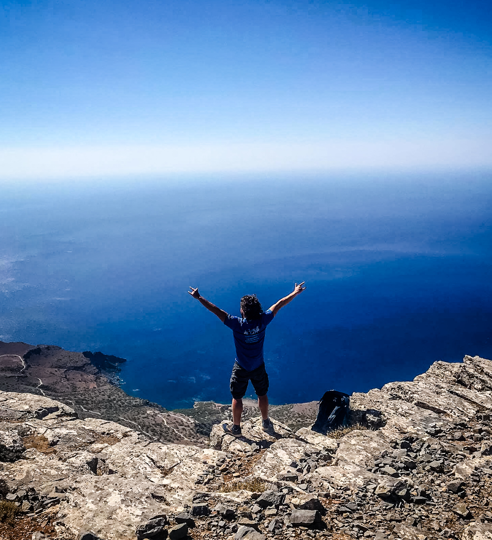 Manolis Zervakis sur un sommet rocheux en Crète, bras levés face à une vue imprenable sur la mer Méditerranée et le ciel bleu.