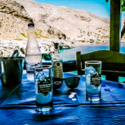 Table au bord de la mer en Crète avec une bouteille d’eau, un verre d’ouzo glacé, et une vue pittoresque sur les montagnes et la mer Égée.