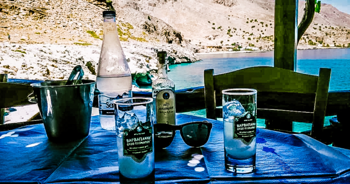 Table au bord de la mer en Crète avec une bouteille d’eau, un verre d’ouzo glacé, et une vue pittoresque sur les montagnes et la mer Égée.