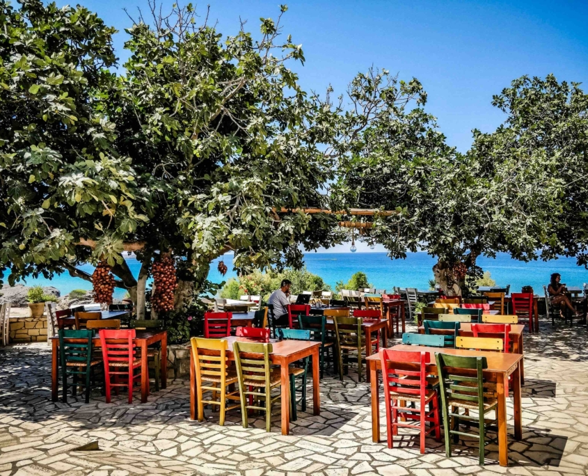 Terrasse colorée d’une taverne traditionnelle en Crète, ombragée par des arbres, avec une vue sur la mer Méditerranée en arrière-plan.